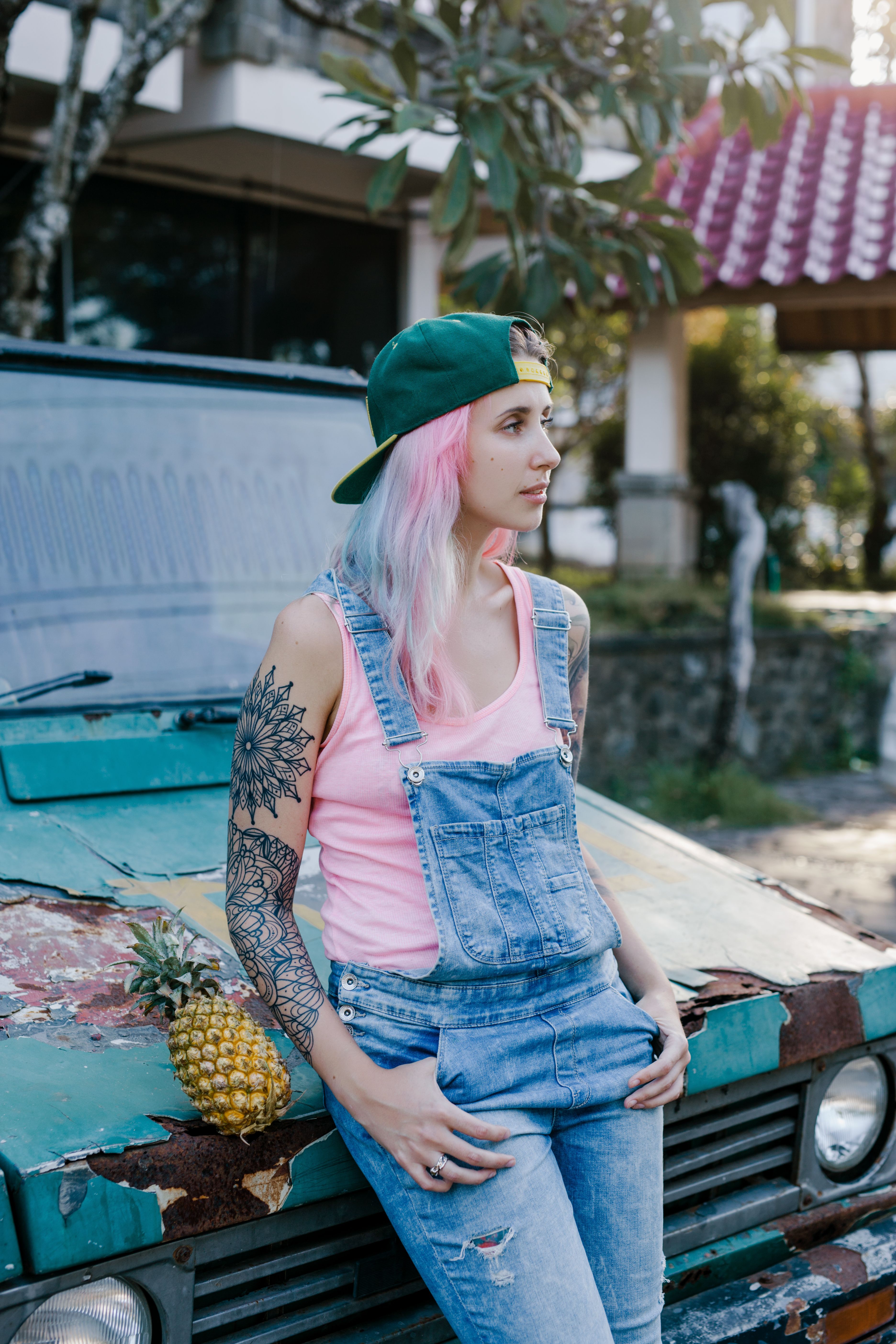 Caucasian woman with pink hair and a tattoo, wearing a denim jumper, leaning on a vintage vehicle with a pineapple.