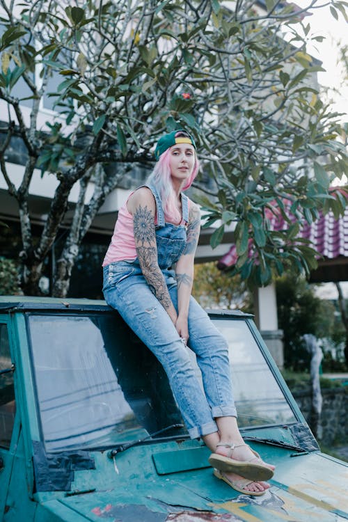 Free Full body positive young female with dyed pink hair and tattooed skin wearing casual denim jumpsuit sitting on shabby old car windshield and looking at camera Stock Photo