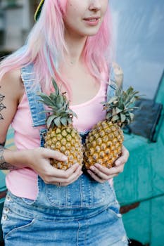 A woman in a denim jumper holding two pineapples, showcasing a vibrant style with pink hair.