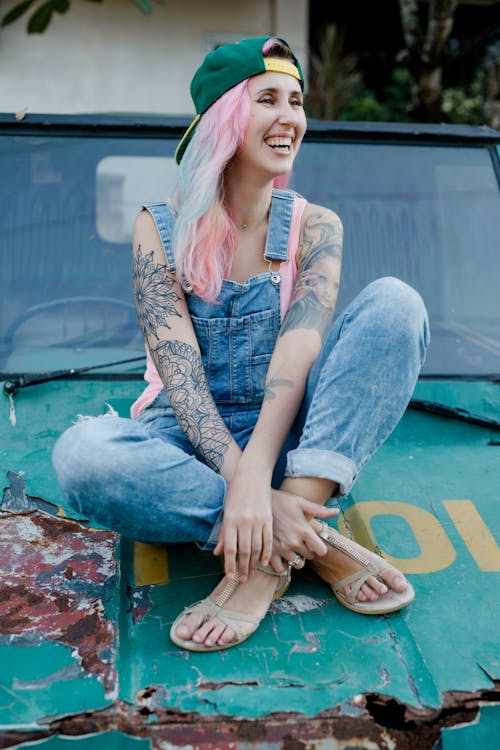 Free Full body smiling informal female with dyed pink hair and tattooed body wearing denim jumpsuit and cap sitting with legs crossed on weathered rusted car hood and looking away happily Stock Photo