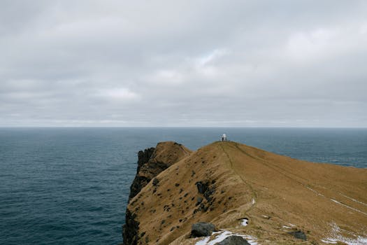 Majestic Icelandic cliff with lighthouse on the edge, overlooking the vast ocean under a cloudy sky.