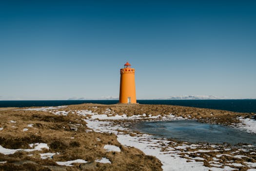 Orange lighthouse on a rocky Icelandic coast with clear blue skies and distant mountains.