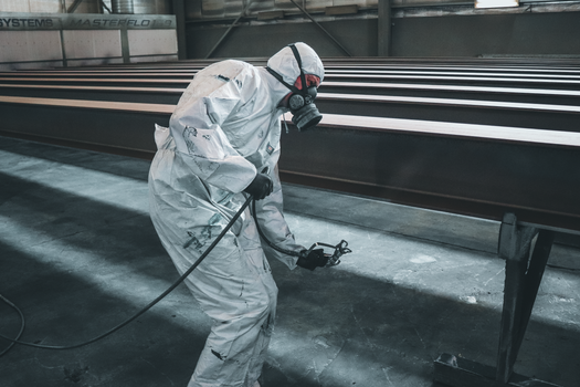 Worker in protective gear spray painting metal beams inside a factory setting.