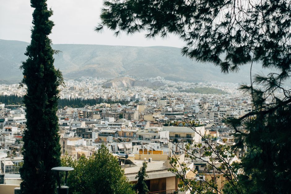 Panoramic view of Athens city buildings with mountains in the background, framed by trees.