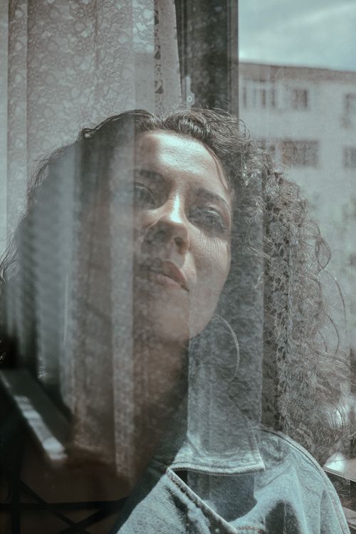 Free Through glass of dreamy female with dark hair looking out window with reflection on glass surface and building on background Stock Photo