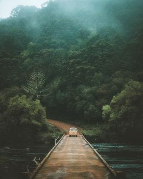 A vintage car travels on a serene bridge through a misty forest in São José dos Ausentes, Brazil.