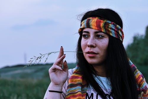 Calm female with dark hair wearing colorful headband while standing in green meadow with blade of grass in mouth on blurred background