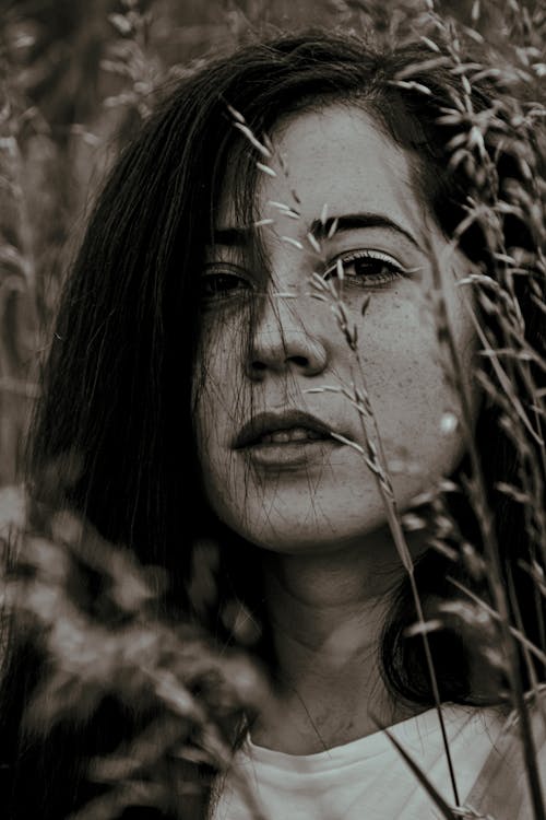 Thoughtful female with dark hair looking at camera while standing near tall grass while resting in nature on summer day