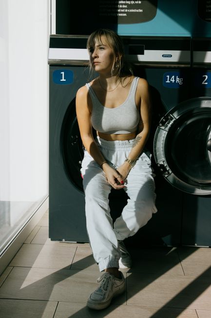 Full body of female in trendy outfit standing in public laundry and looking away at sunlight