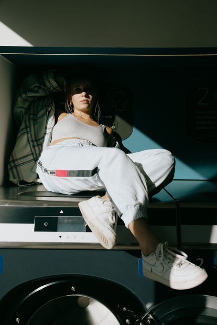 Full body of serious female in trendy clothes hiding face from sunlight in shade while resting on washing machine and looking at camera