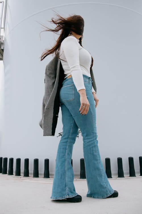 From below of plump female with long hair waving in wind wearing jeans with white sweater standing against white wall