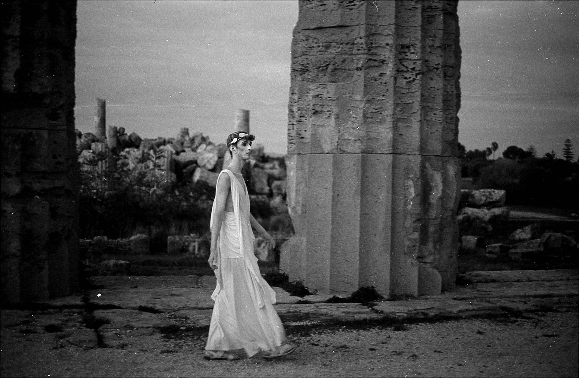 Grayscale Photo of a Man in Ancient Greek Clothing Walking