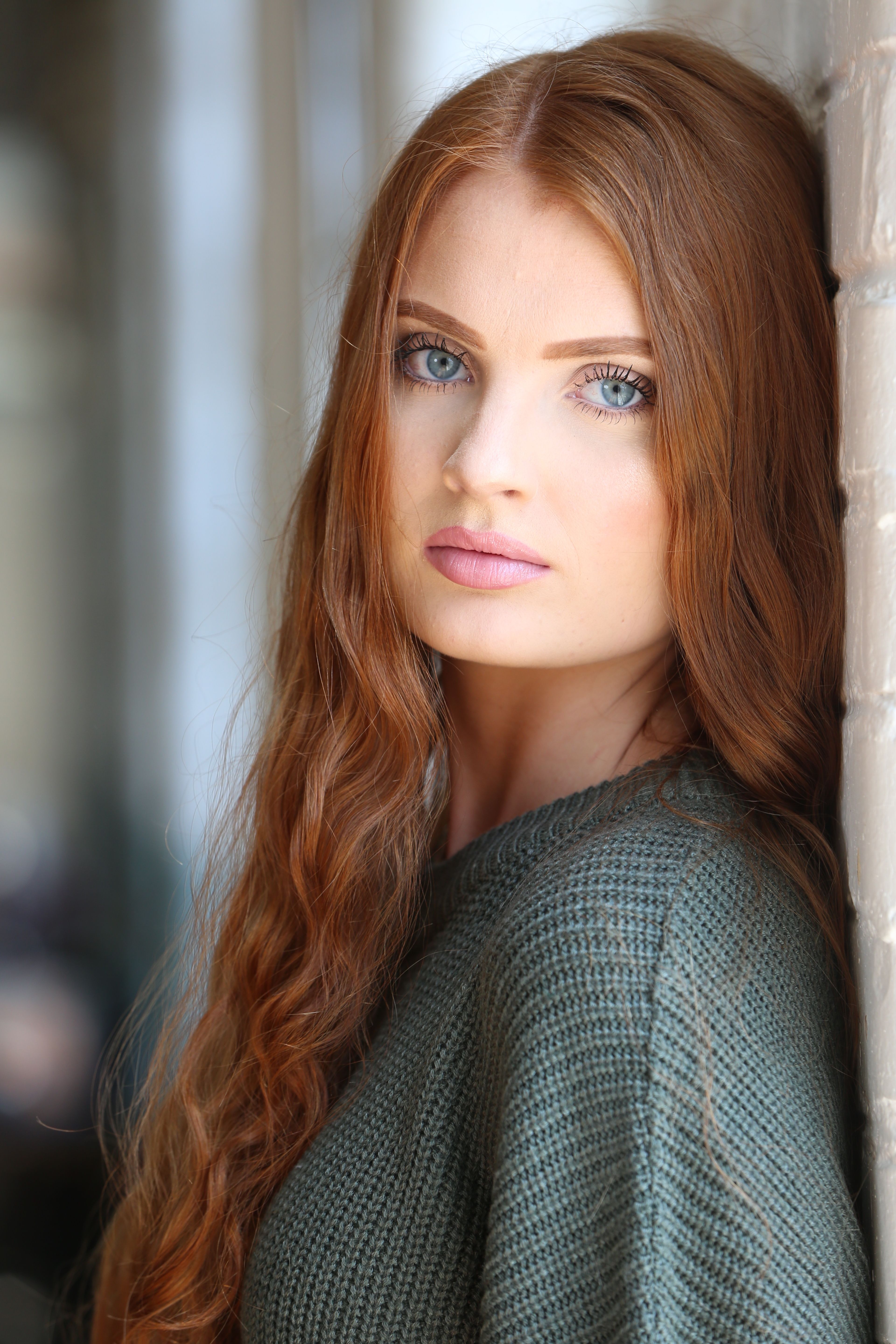 Elegant portrait of a young woman with long red hair leaning against a white wall.
