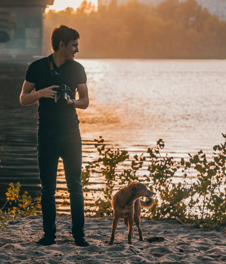 Man With Dog And Camera Near River At Sunset