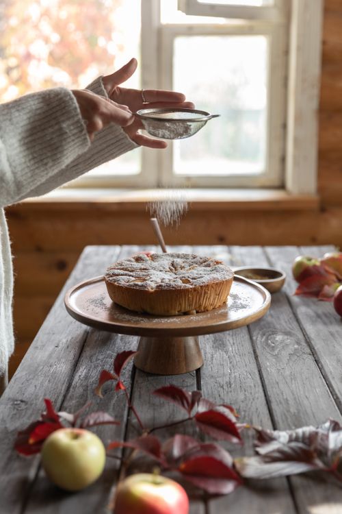 Free A cozy apple cake being dusted with powdered sugar on a wooden table in natural light. Stock Photo