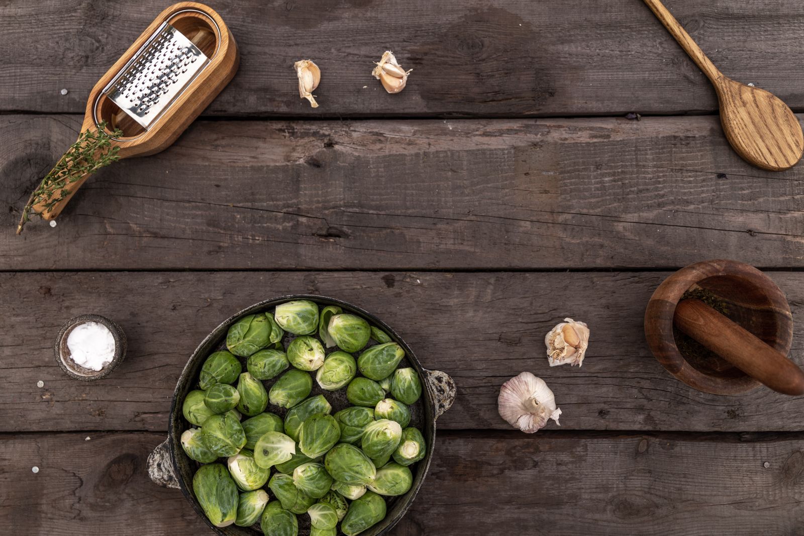 Overhead image of Brussels sprouts and garlic on a rustic wooden surface with kitchen tools.