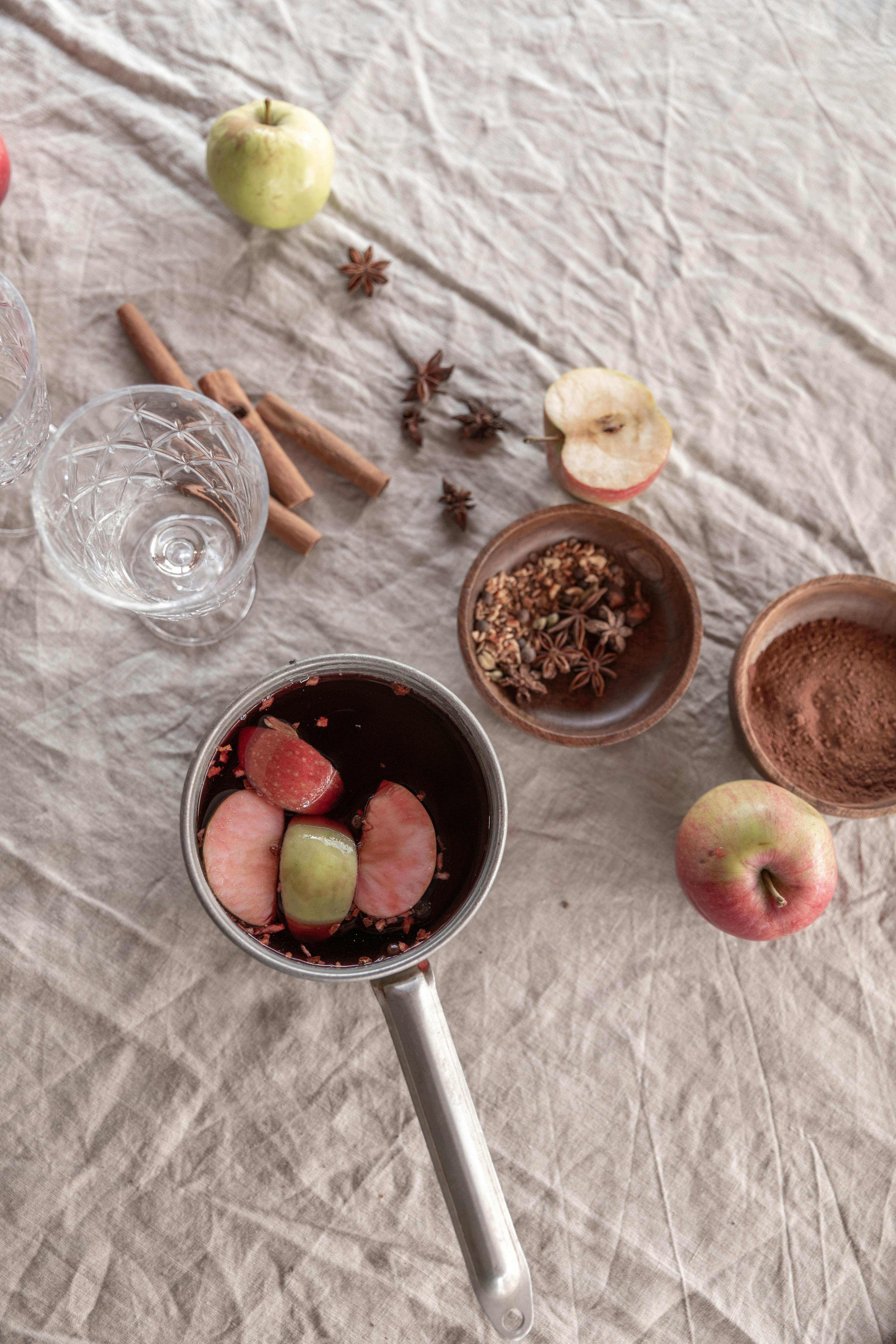 Overhead view of mulled wine preparation ingredients with apples, spices, and cookware on a linen tablecloth.
