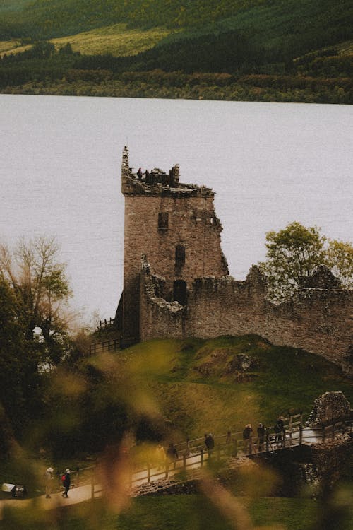 From above of aged partially destroyed castle facade on green mount near bridge with unrecognizable travelers