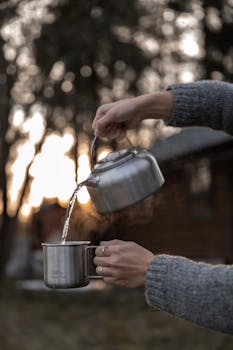 A person pours hot water from a kettle into a cup outdoors at sunrise, creating a warm, relaxing atmosphere.
