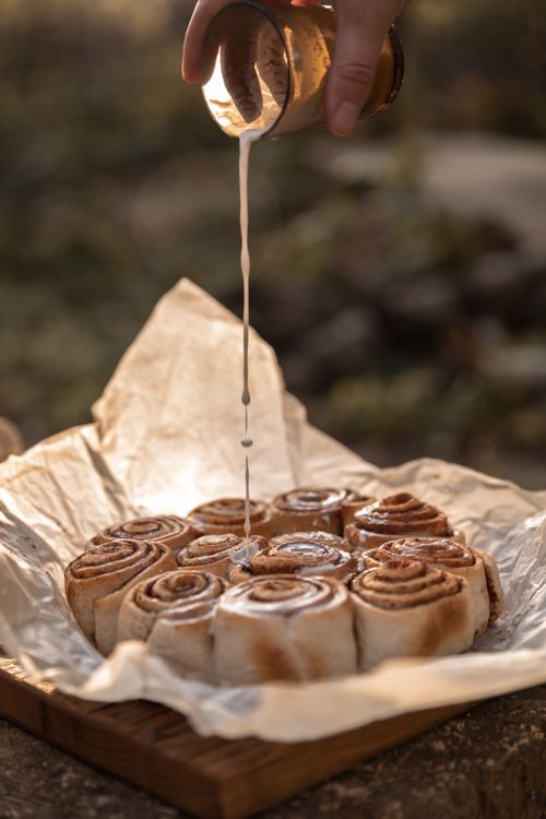 Free Warm cinnamon rolls being drizzled with icing in an outdoor setting on parchment paper. Stock Photo