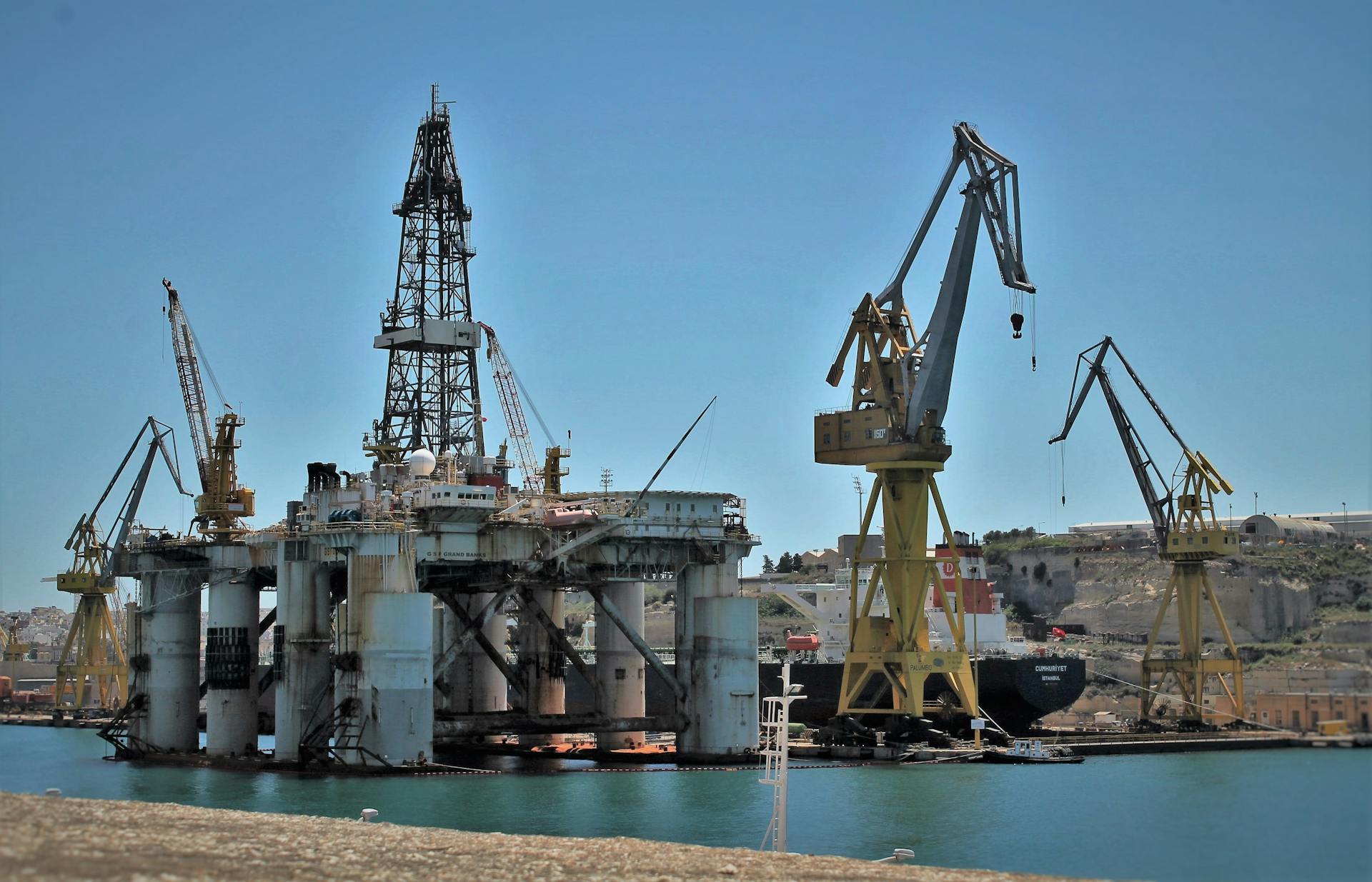 An offshore oil rig surrounded by cranes in a sunny industrial harbor setting.