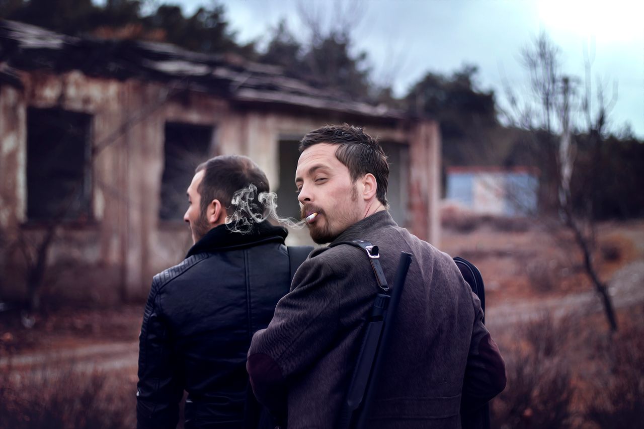 two men in military style attire smoking outdoors near abandoned build