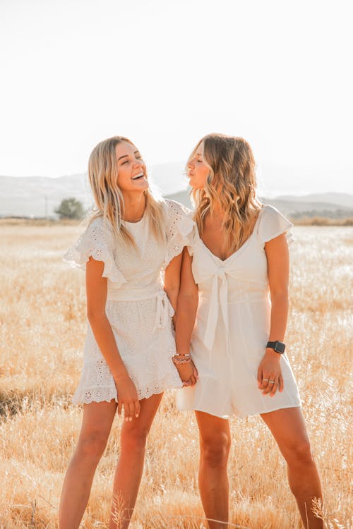 Young cheerful female holding hand of best friend with pouting lips on grass meadow under white sky in back lit