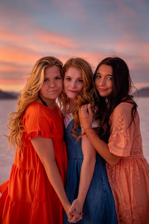 Young cheerful women in bright trendy apparel looking at camera under cloudy sky at sundown