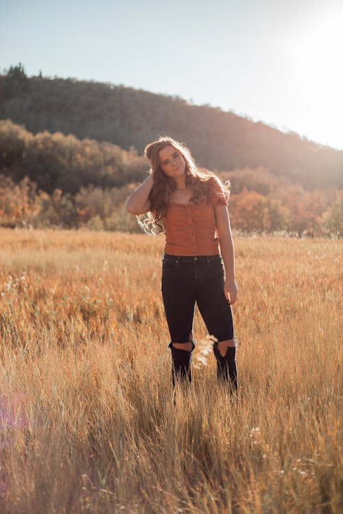 Young female in trendy apparel with hairstyle standing in field behind mountain while looking at camera in back lit