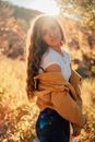 Side view young pretty female with long curly hair lowering jacket and looking at camera while standing in sunny autumn nature
