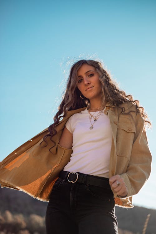 From below content young female with long curly hair wearing stylish jacket standing on sunny nature against cloudless blue sky and looking at camera