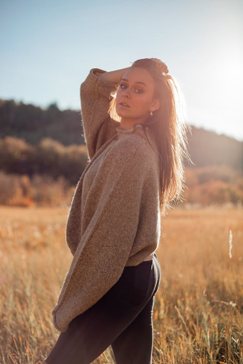 Side view slim elegant female wearing stylish loose sweater standing on spacious field and touching head while looking at camera on sunny autumn day