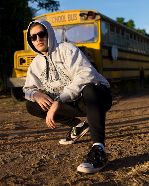 Serious young male in casual outfit and sunglasses sitting on hunker near yellow school bus on terrain under blue cloudless sky near trees in daylight