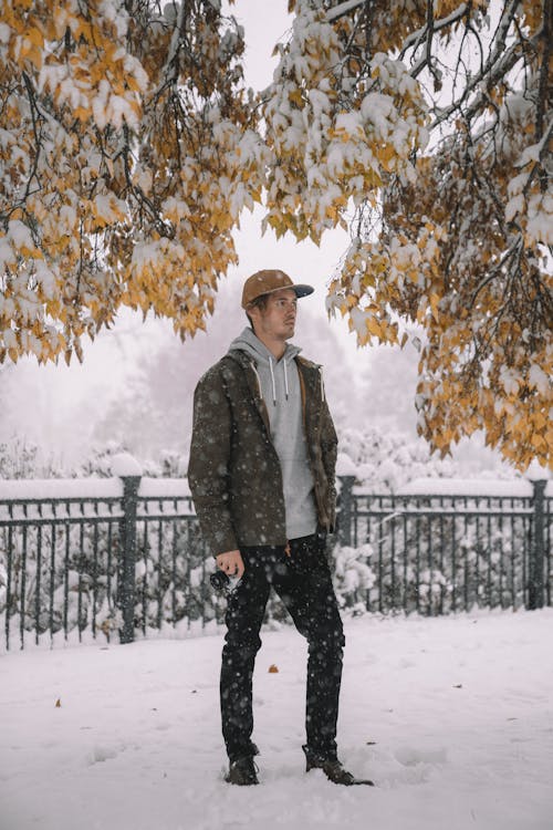 Full body man in cap and jacket holding photo camera while standing at fence in snowfall