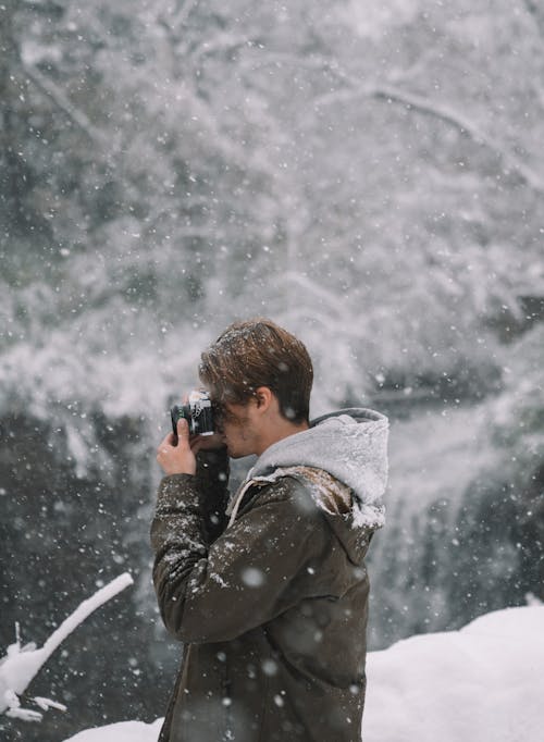 Side view of hipster guy with photo camera taking shot of snowy landscape in nature