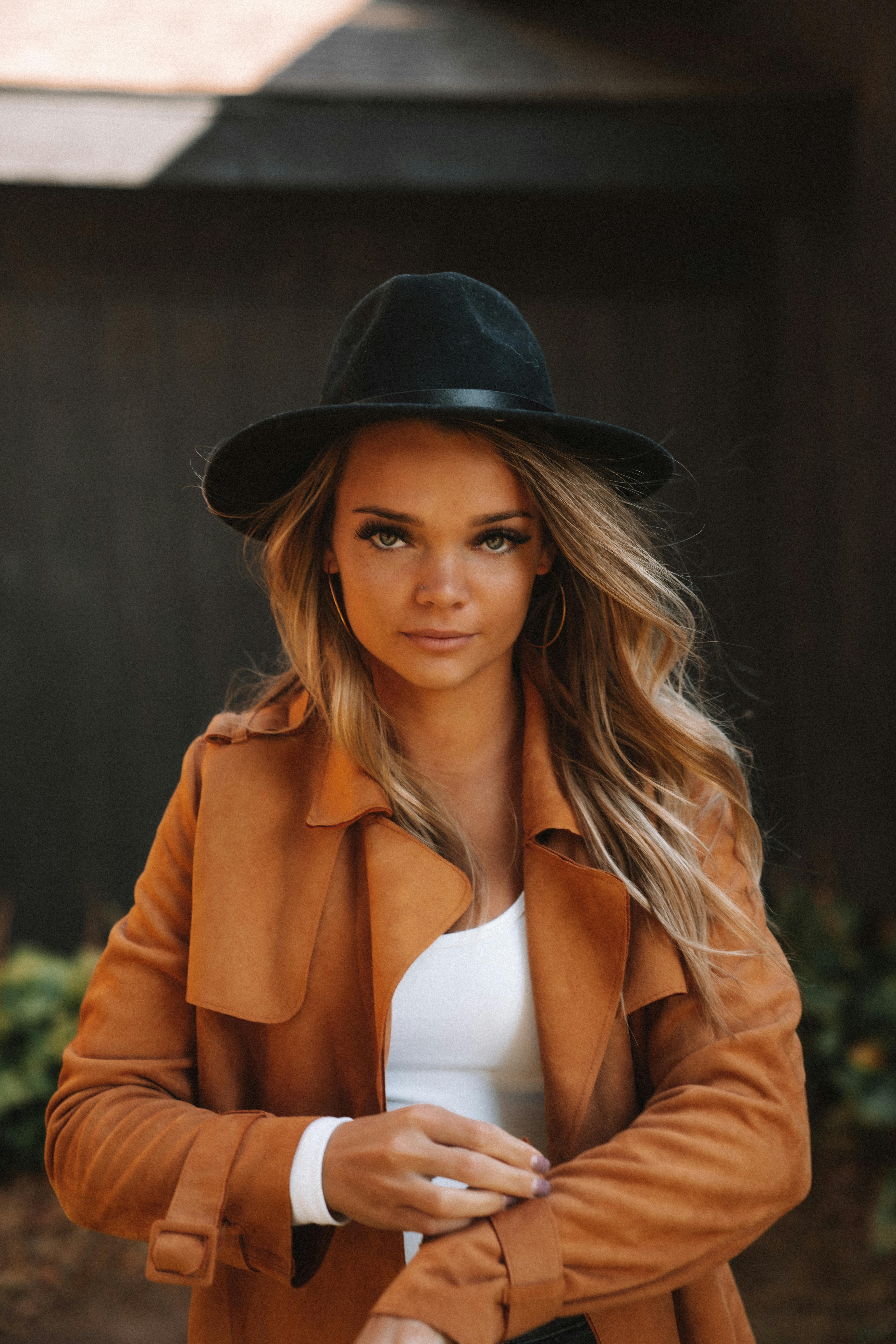 Young female in trendy coat and hat looking at camera while standing on blurred background of city street