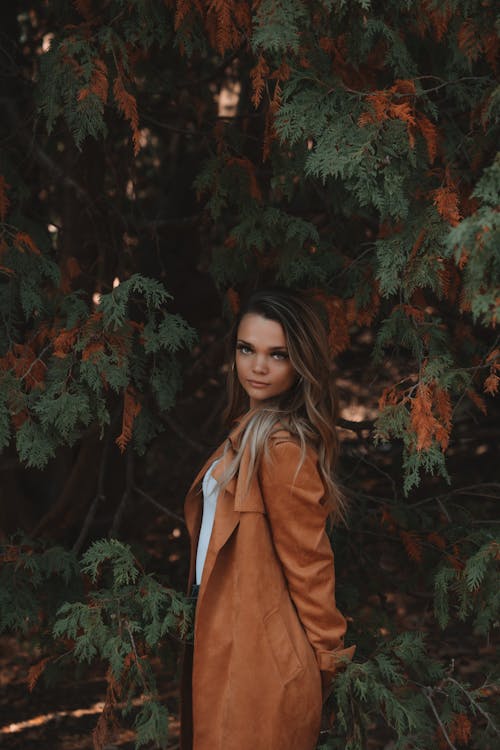 Young confident woman in stylish coat standing against trees in autumn time looking at camera