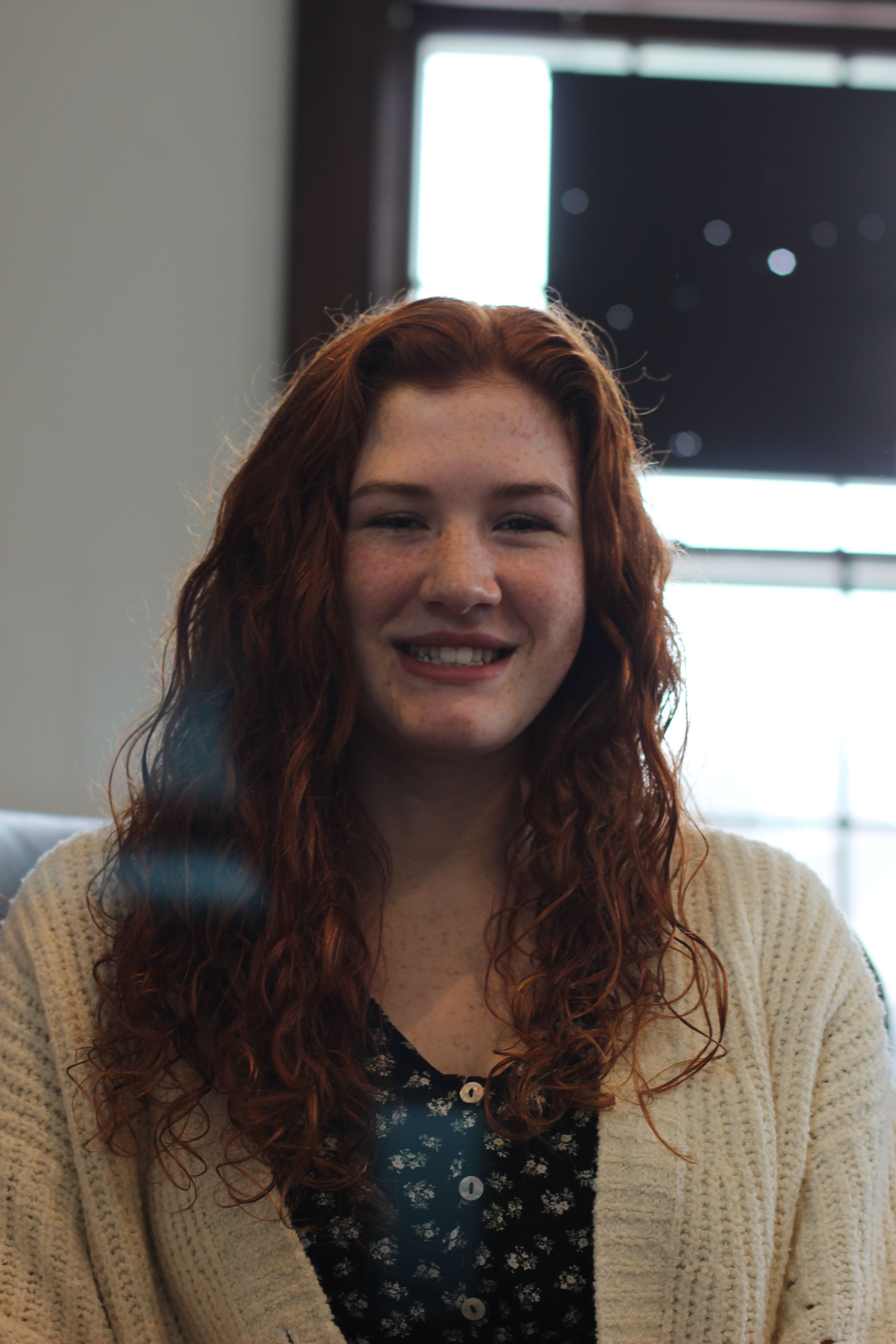 Portrait of a smiling young woman with red curly hair and a cozy sweater indoors.
