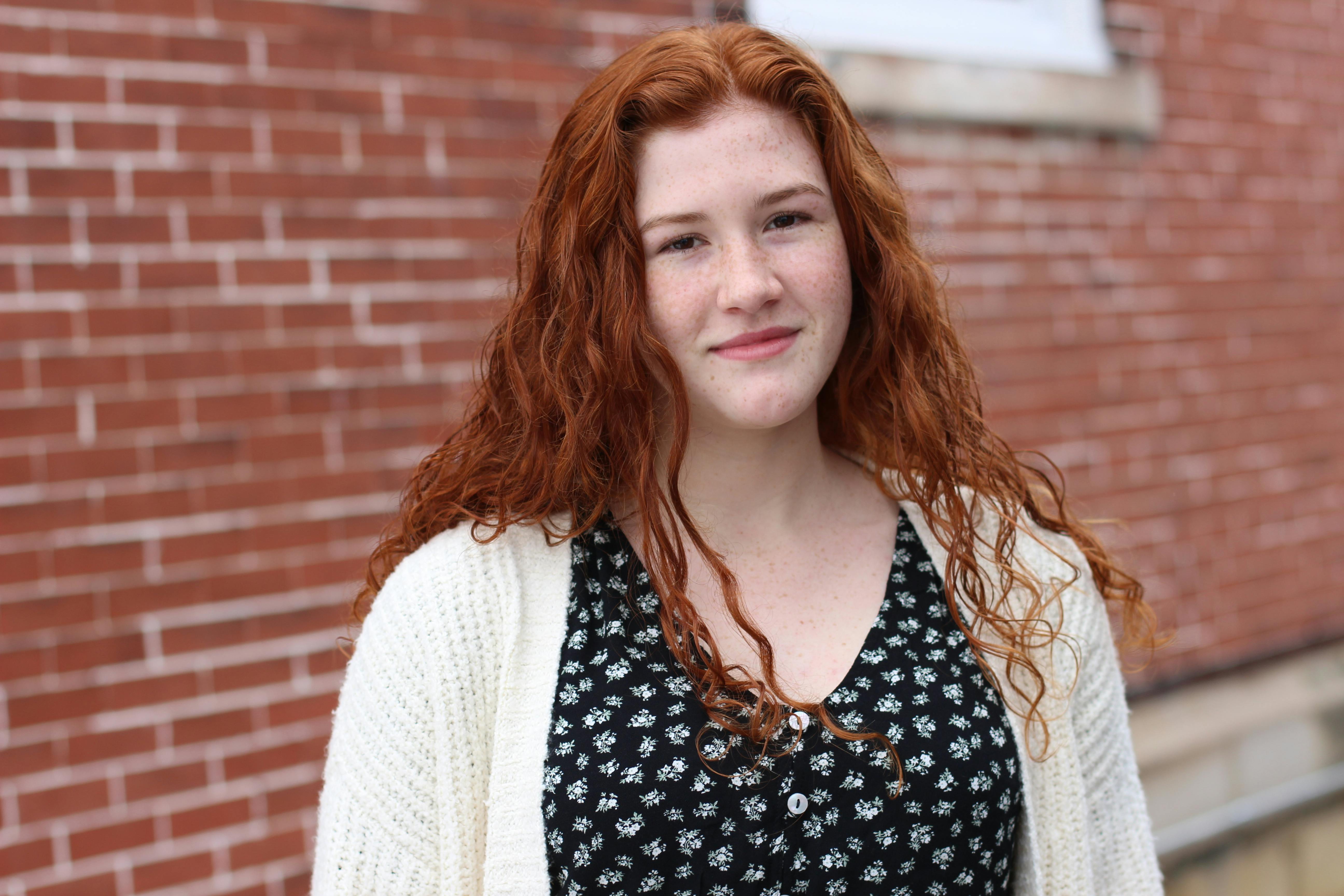 Portrait of a smiling woman with curly red hair and freckles standing by a brick wall.