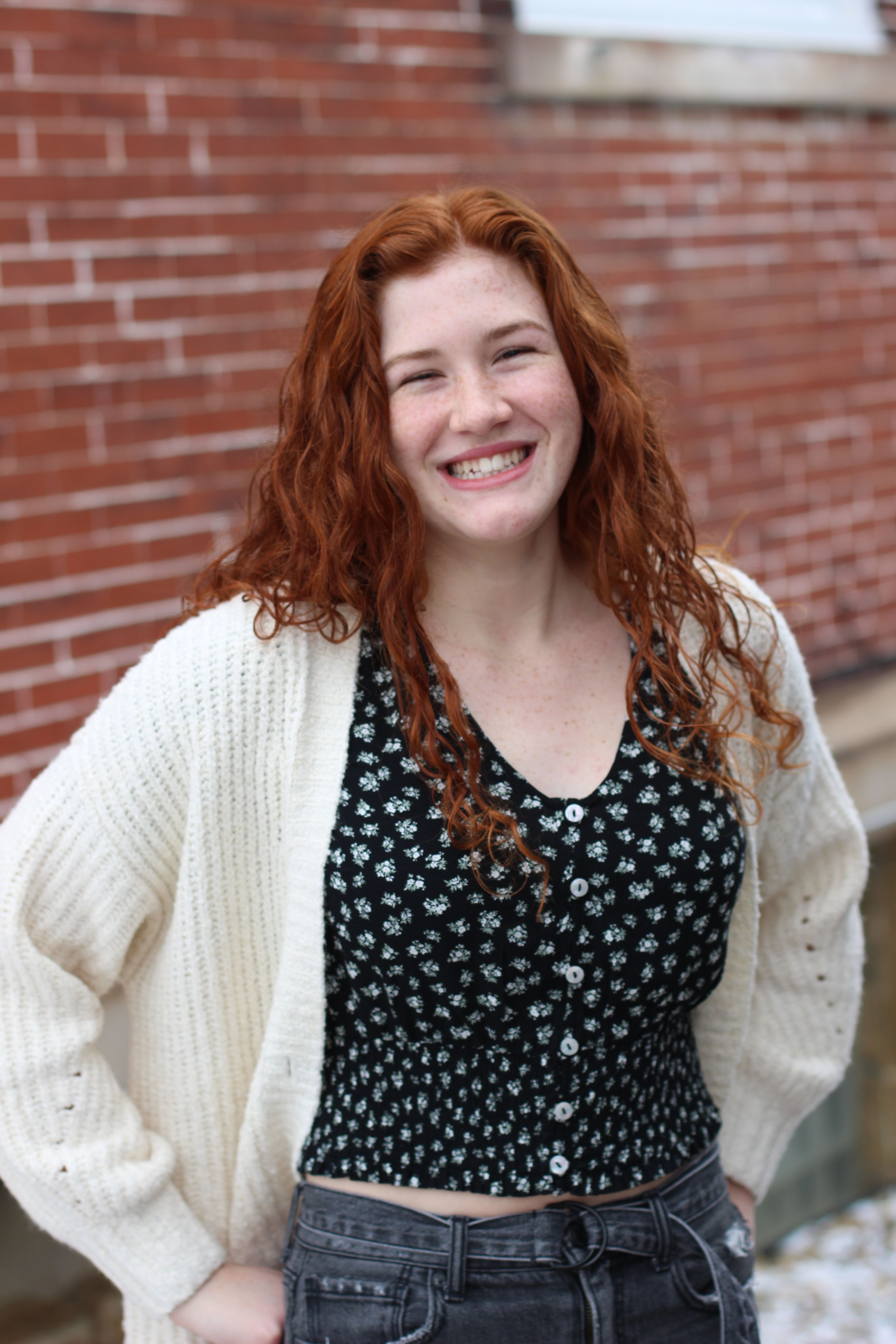 Smiling woman with red curly hair in cardigan standing against a brick wall.