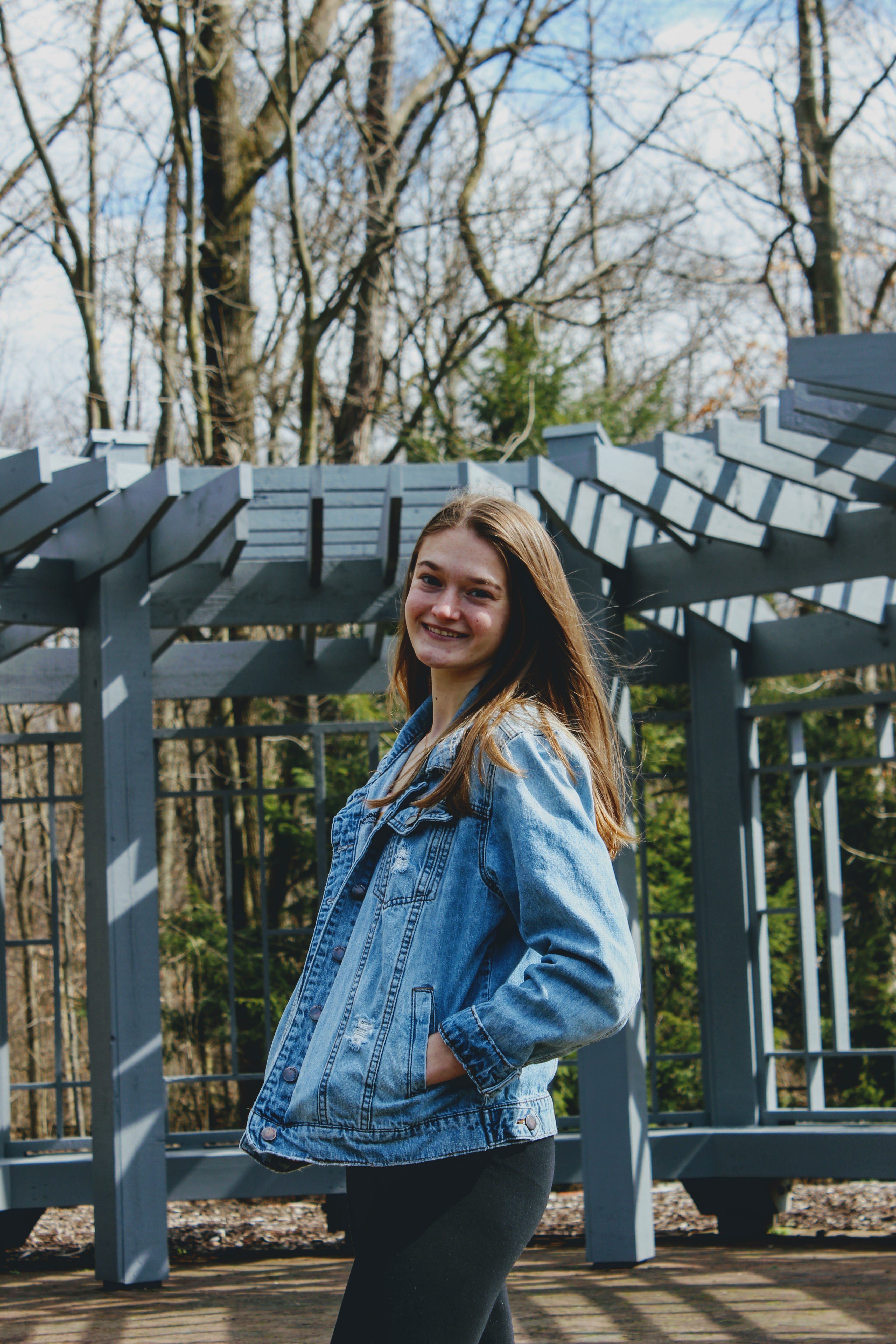Portrait of a smiling young woman posing in a denim jacket outdoors with trees and structures in the background.
