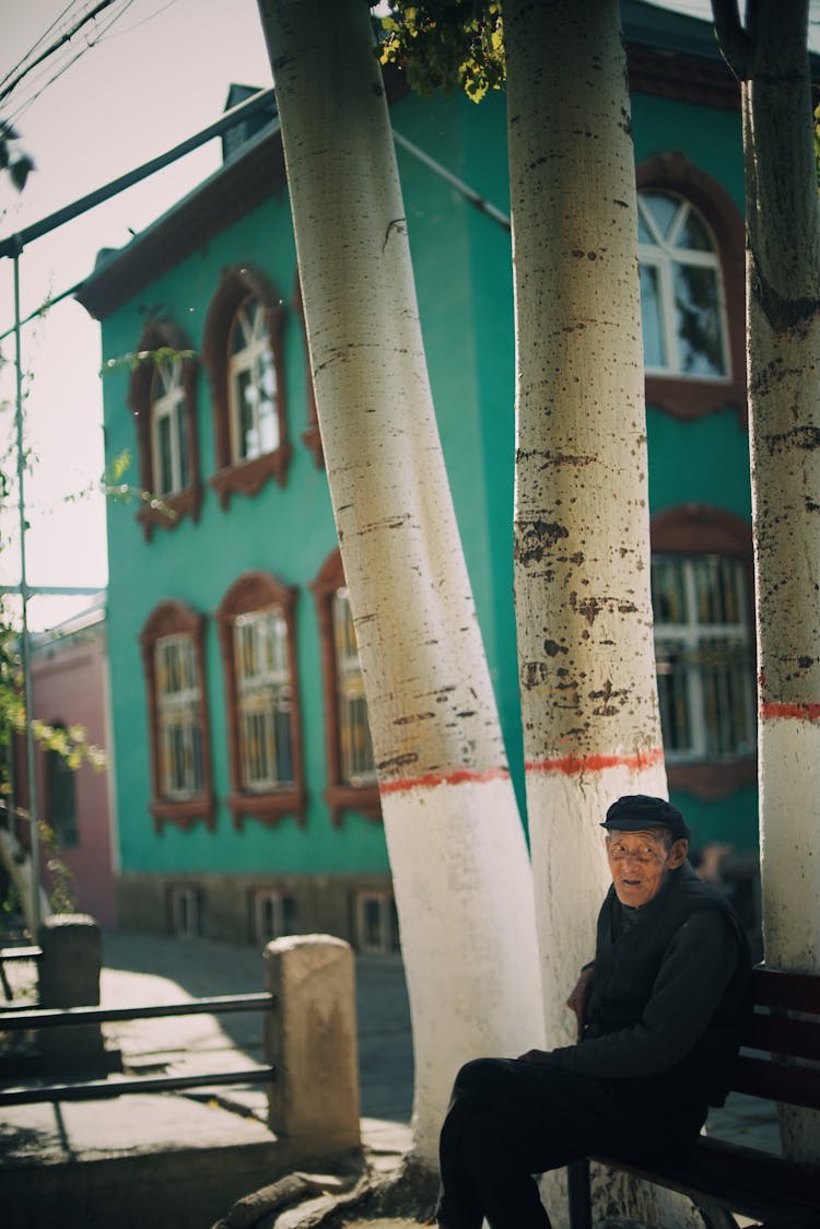 Senior Man Sitting By White Painted Tree Trunks, And Turquoise House In Background