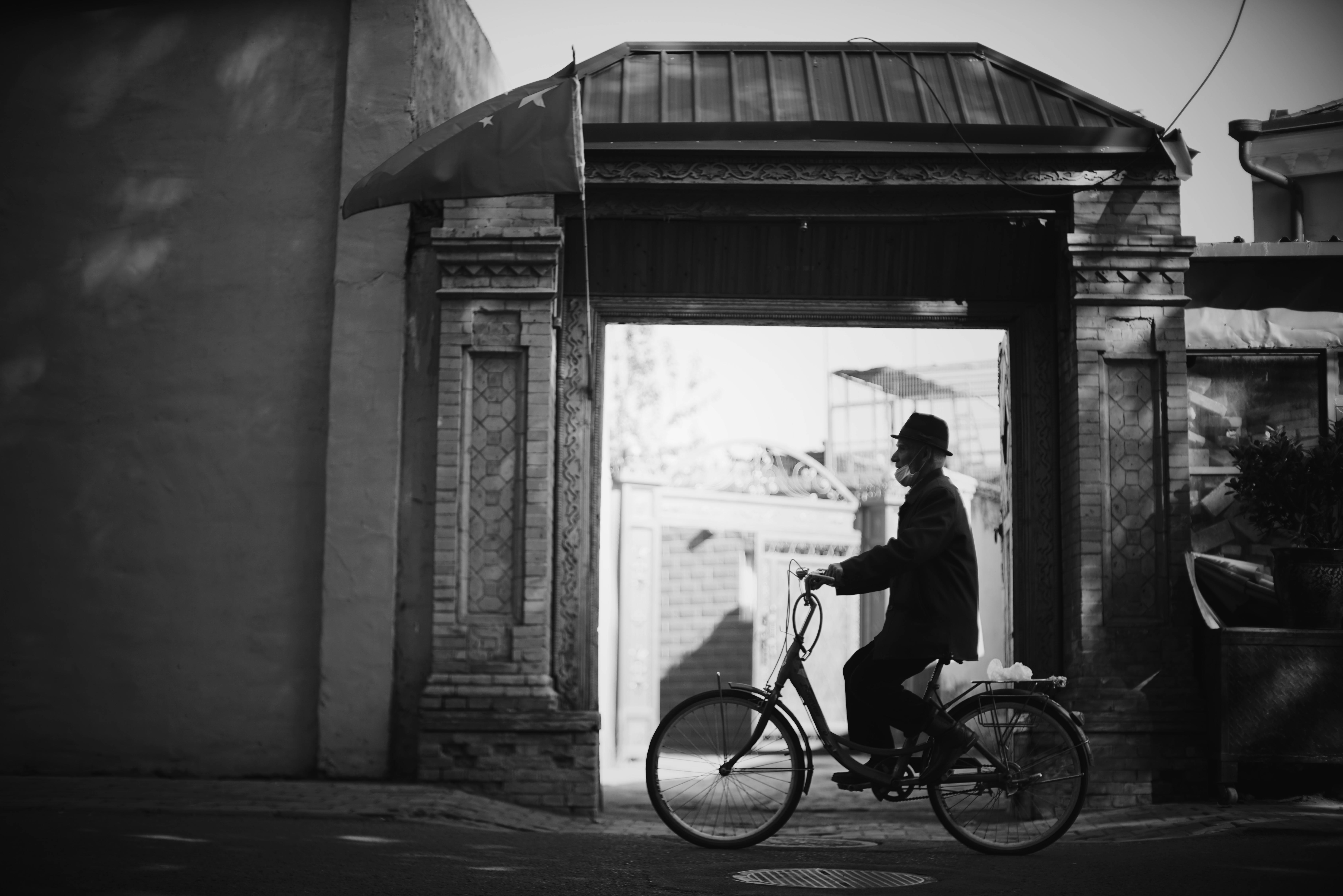A black and white image of an elderly man cycling through a large, ornate archway, wearing a hat.