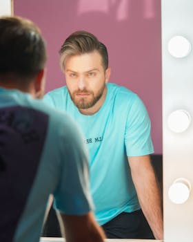 Portrait of a man with a beard looking at his reflection in a well-lit makeup mirror.