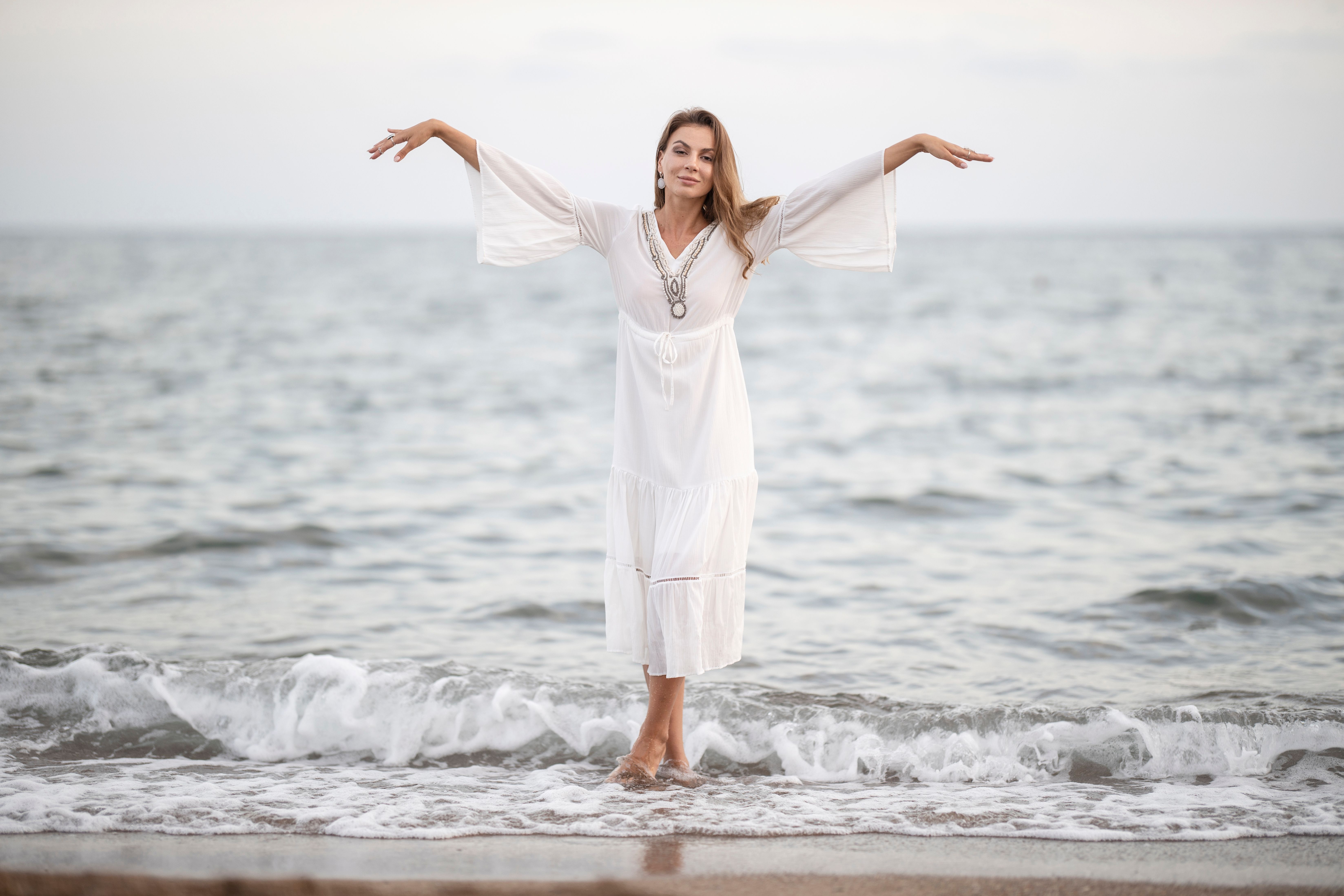 Elegant woman in white dress standing at the seaside, embracing the serene ocean backdrop.