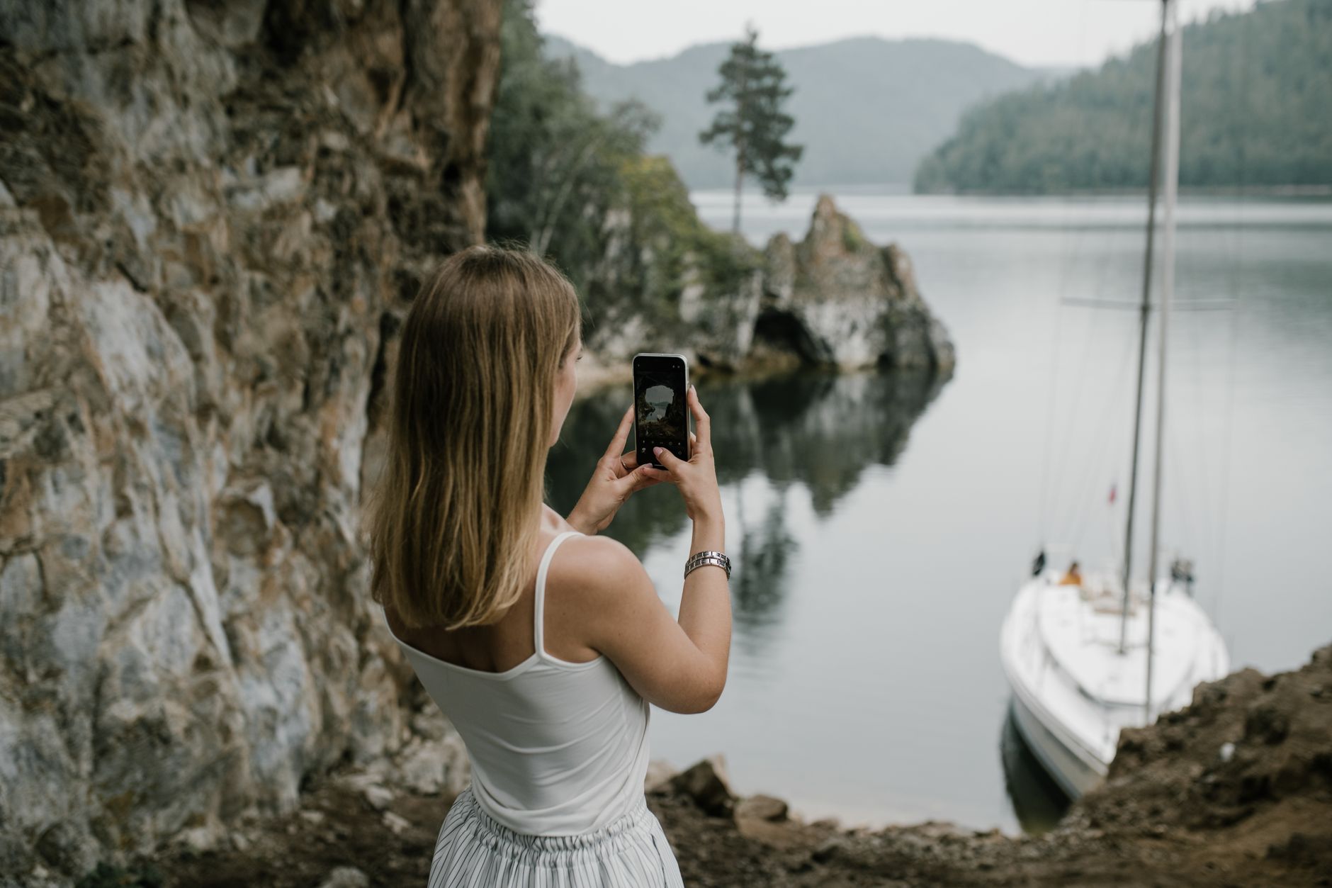 新手手機攝影設定表:白天夜景室內一張圖速查 A woman takes a photo of a serene lakeside with a sailboat in lush natural surroundings.