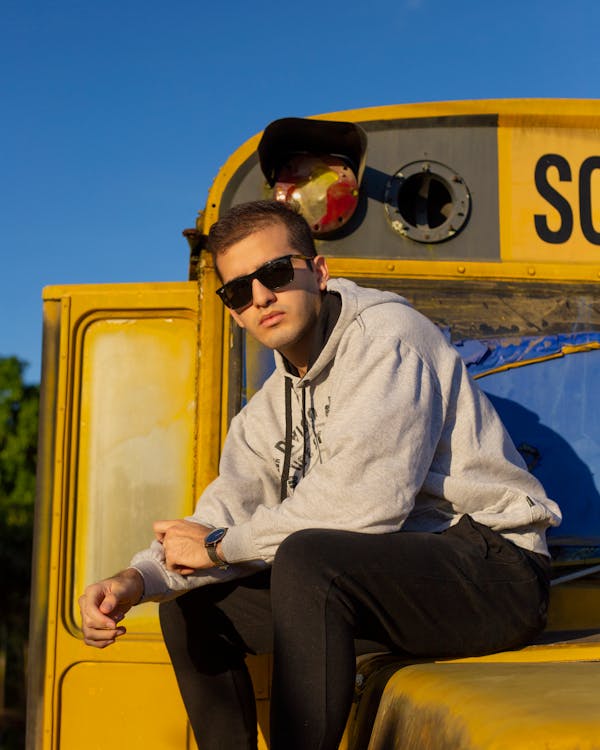 Self assured young male traveler in trendy outfit and sunglasses sitting on yellow bus and looking at camera against bright blue sky on sunny day