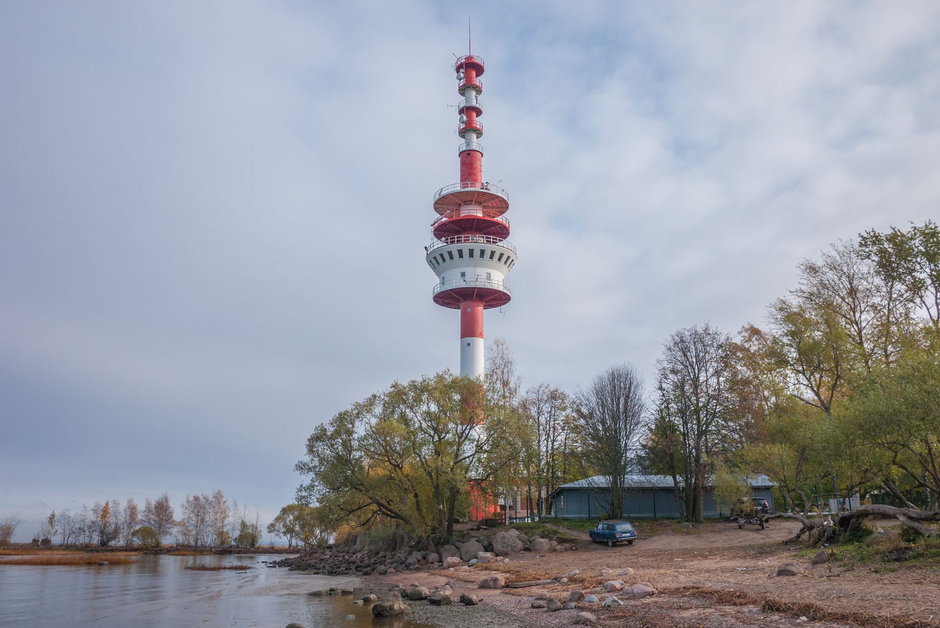 Landscape with towering red and white communications tower beside a lake and trees.