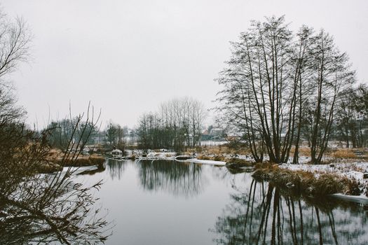 A serene winter landscape featuring bare trees reflected in a calm lake under a cloudy sky.