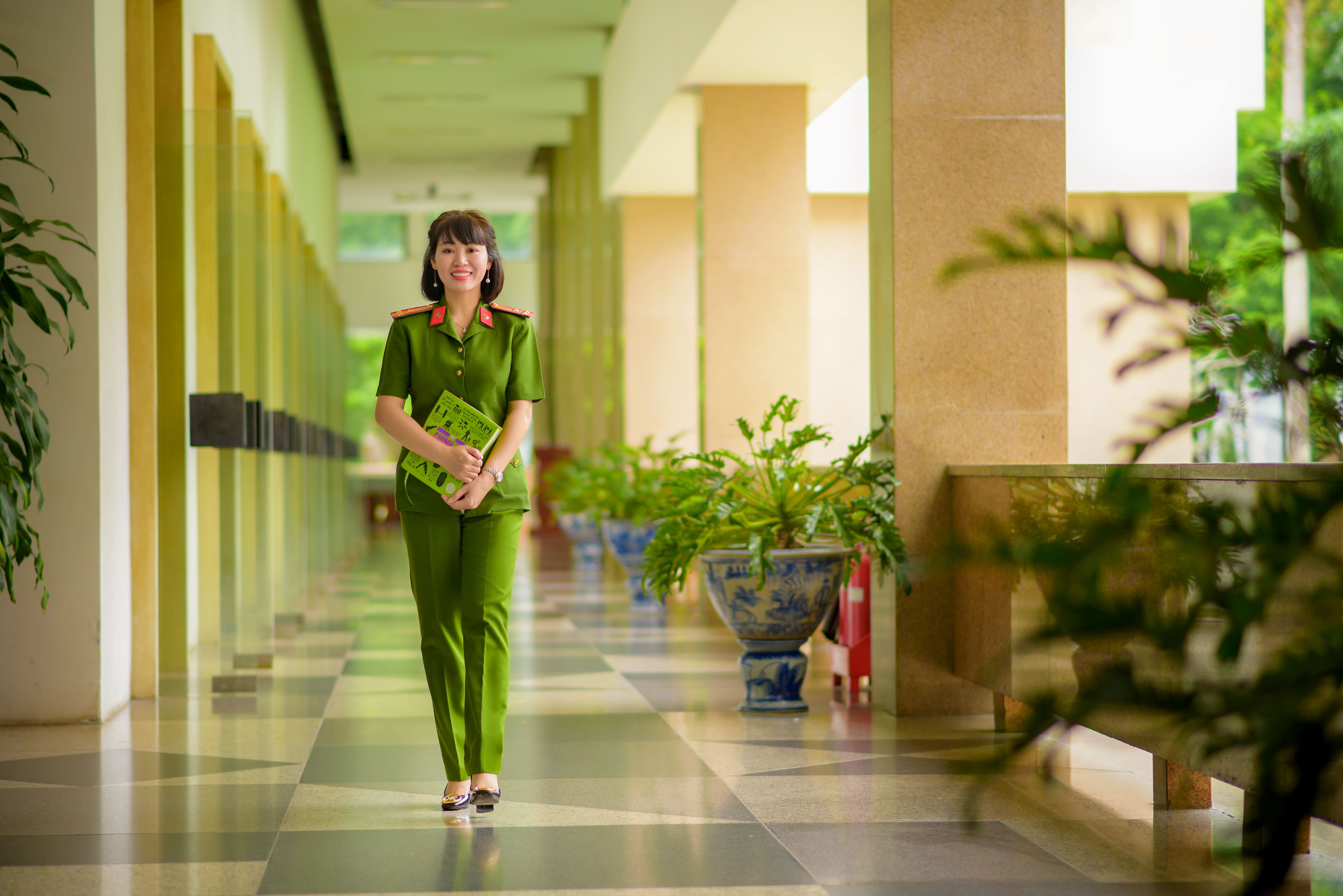 Asian woman in green military uniform walks through a bright hallway, holding a book and smiling.